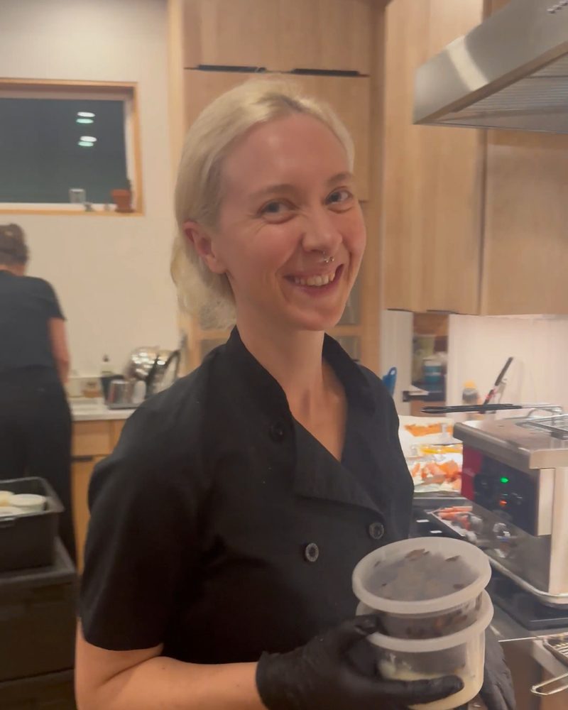 Chef Mollie in a black chef's jacket, smiling in a client's kitchen while her team works behind her during a dinner service