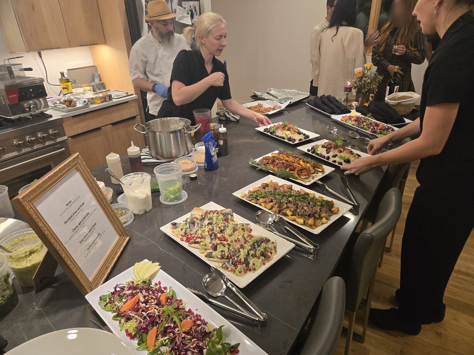 A family-style private dinner by Chef Mollie set out on a client's kitchen island: bronzino with salsa verde, slow-roasted za'atar lamb, honey-harissa squash, jeweled rice and a chicories salad, with Chef Mollie plating and a framed menu at the end of the counter